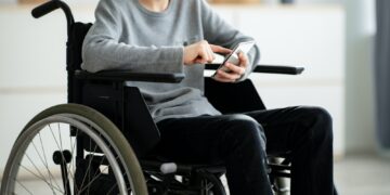 Cropped view of disabled teen boy in wheelchair using smartphone at home, closeup