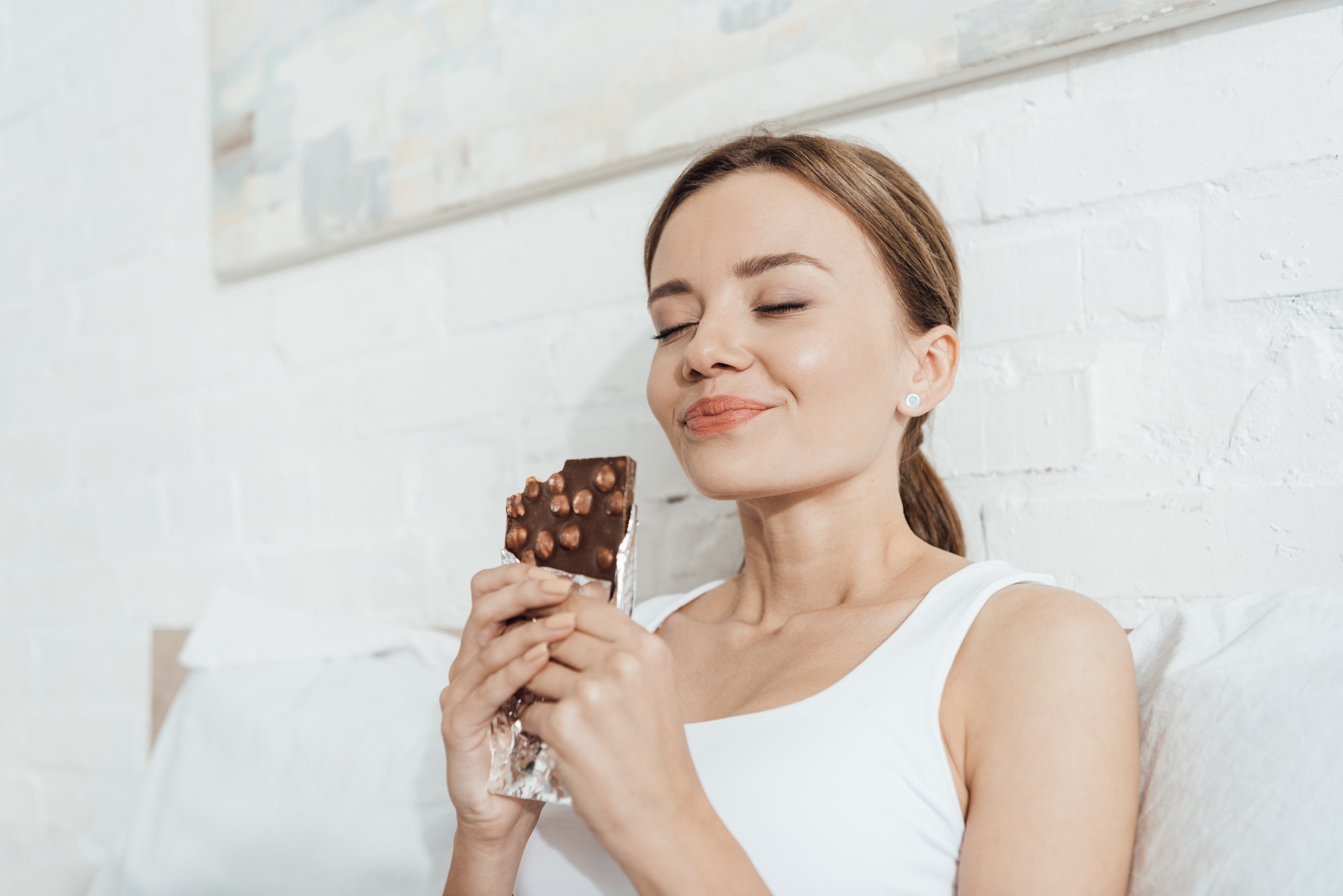 low angle view of smiling young woman eating chocolate with nuts with closed eyes in bed