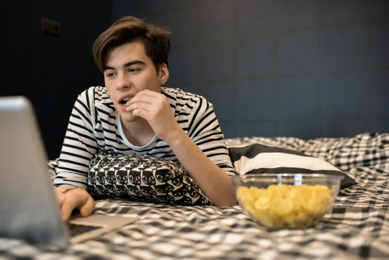 Young man eating chips using laptop in bed