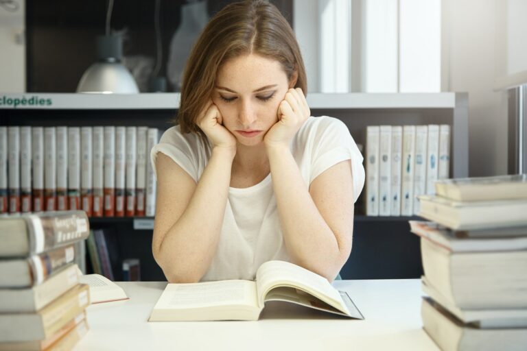 Bored student girl reading a book holding her head in hands, looking concentrated, preparing for the