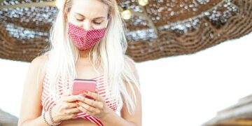 Happy young woman using mobile phone with face mask in beach - Teenager watching video in internet