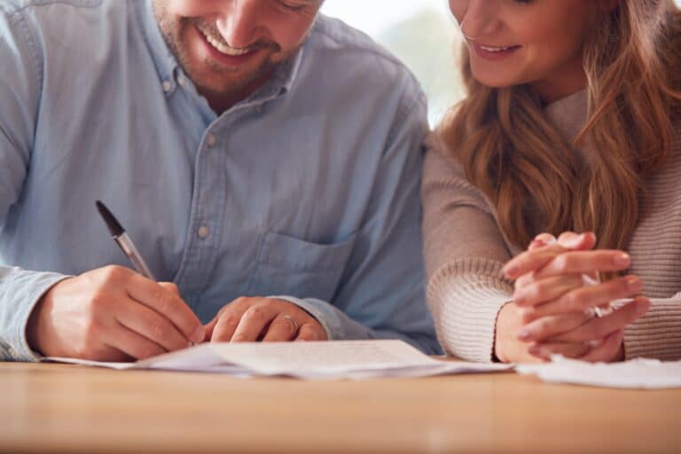 Close Up Of Smiling Couple Signing Financial Document At Home