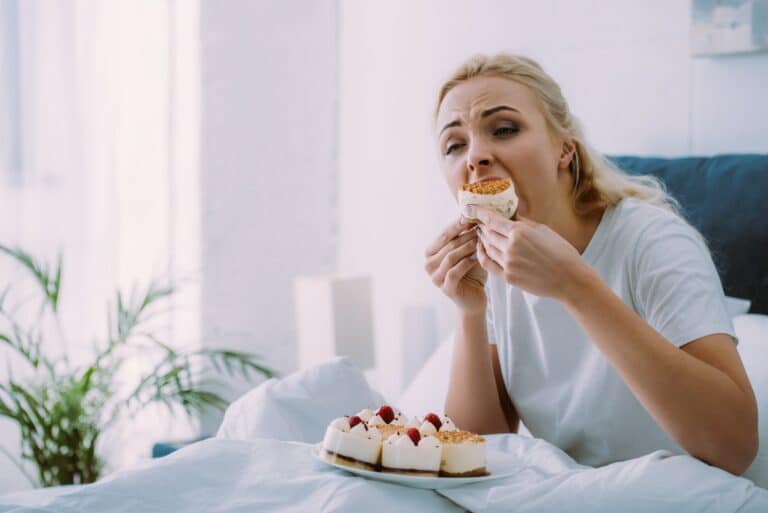 selective focus of sad woman in pajamas eating cake in bed alone