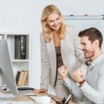 smiling female mentor looking at happy young businessman working with desktop computer in office