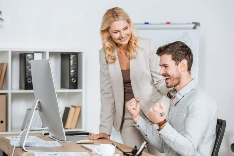 smiling female mentor looking at happy young businessman working with desktop computer in office