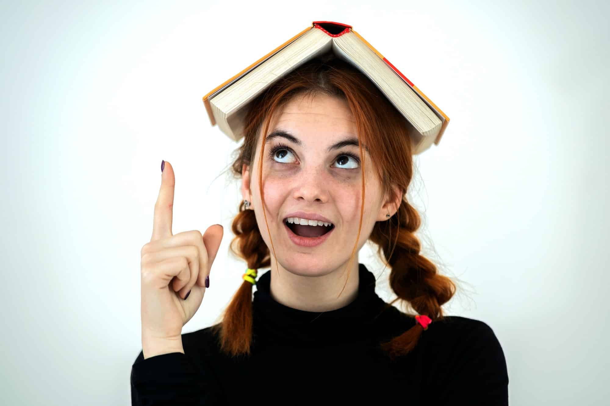 Portrait of funny young smiling student girl with an open book on her head holding her point finger
