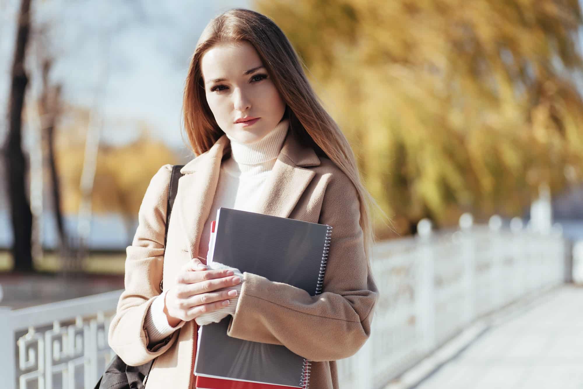blonde student girl stands in the autumn park with notepad in hands
