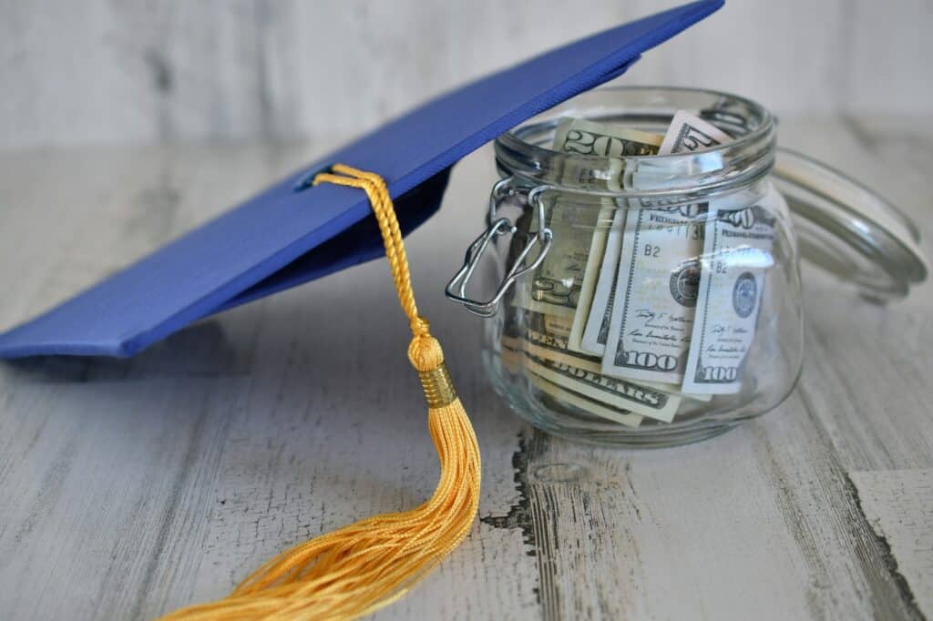 Graduation cap mortarboard with tassel propped on a jar of money cash, concept school loans debt