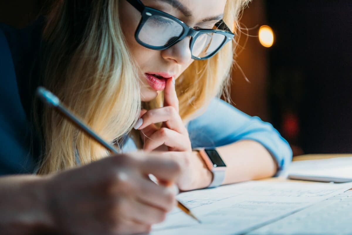 young blonde caucasian student in eyeglasses thinking and writing something with pencil studying e1656006247824