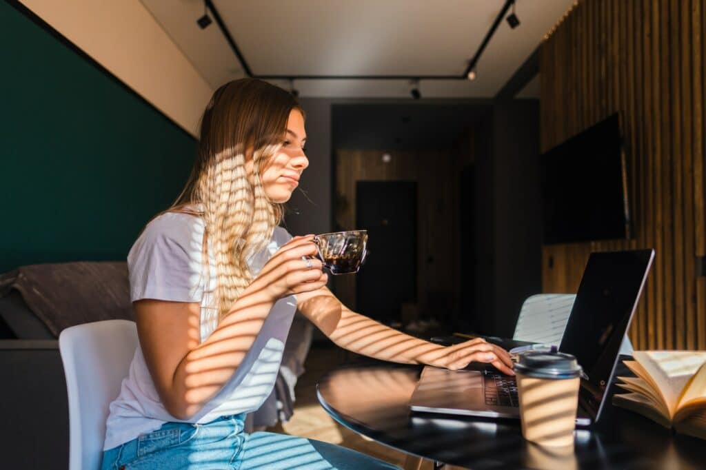 young caucasian woman drinking coffee in the kitchen working with laptop on sunny morning