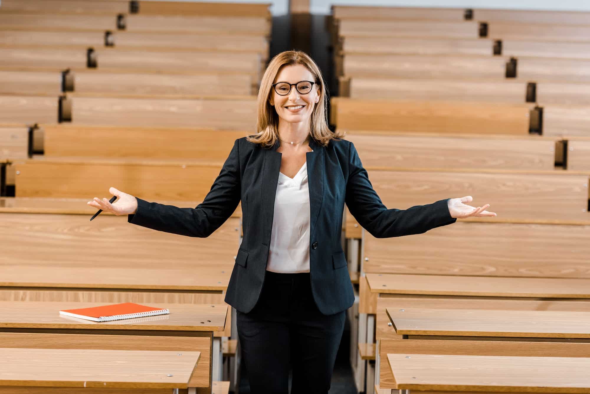 happy female university professor with outstretched hands looking at camera in classroom