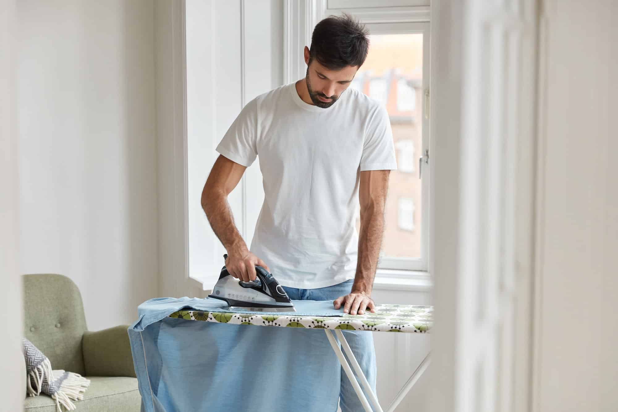 photo of busy unshaven man irons shirt on ironing board prepares for formal meeting on business con