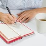 The hand of a woman with a pen writes in her diary. Next on the table is a cup of tea