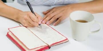 The hand of a woman with a pen writes in her diary. Next on the table is a cup of tea