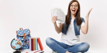 young excited woman student holding bundle lots dollars cash money spread hands sitting near globe backpack school books isolated