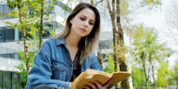 Young student girl in denim jacket reading a book at alley near modern building of University.
