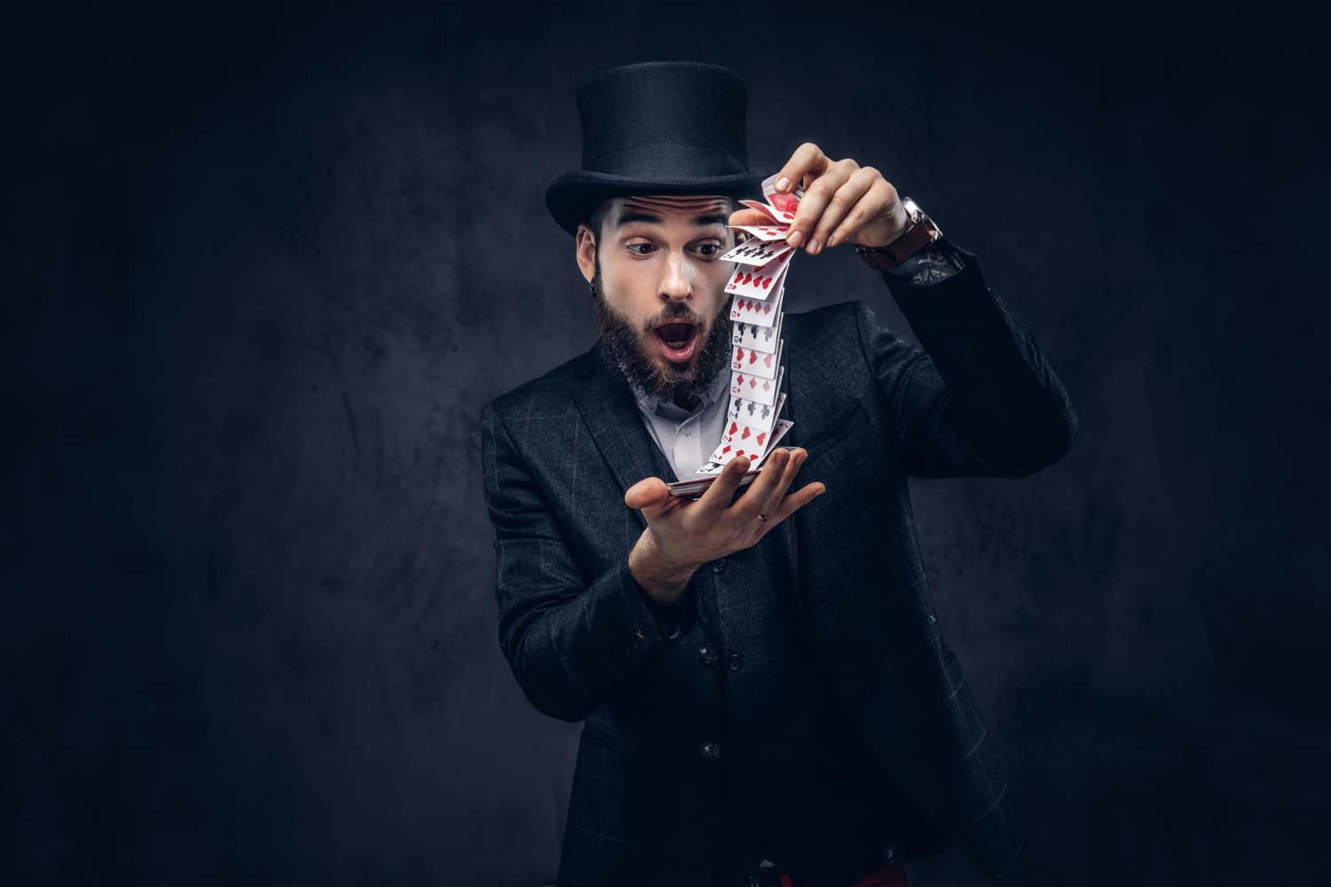 bearded magician black suit top hat showing trick with playing cards dark background