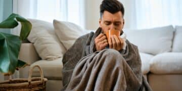 Sick man wrapped in blanket drinking tea at home.