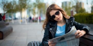 Young woman sitting on bench among urban space and reading newspaper
