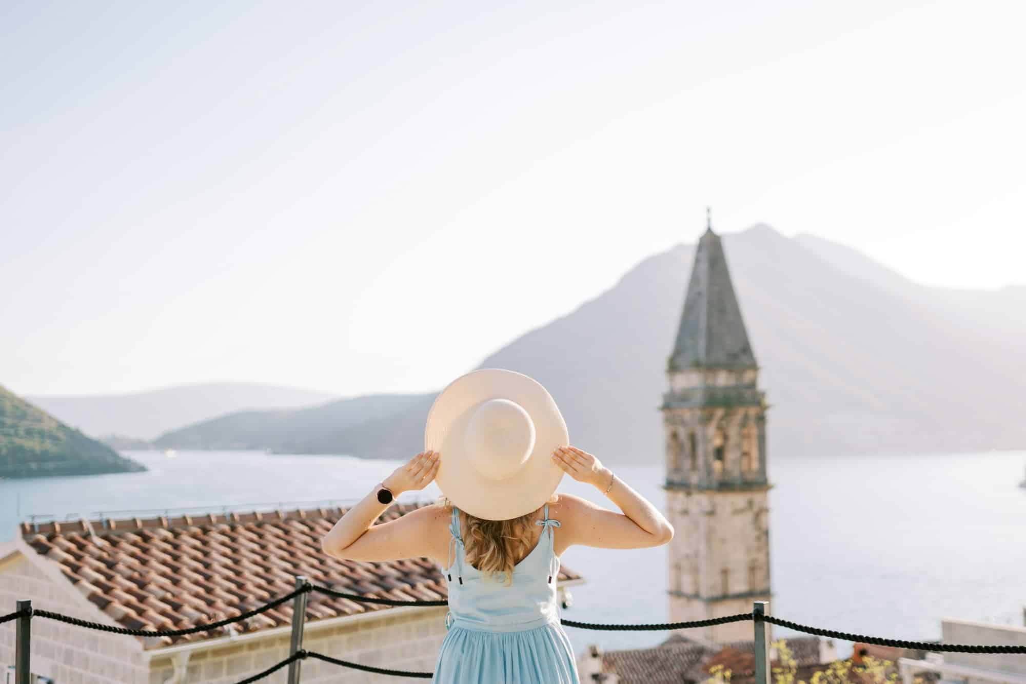 girl stands on the observation deck near the bell tower holding her hat with her hands perast