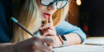 young blonde caucasian student in eyeglasses thinking and writing something with pencil studying