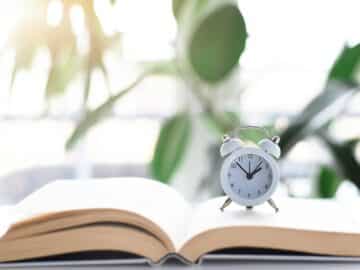 clock on top of an open book with blurry green houseplants in the background reading time